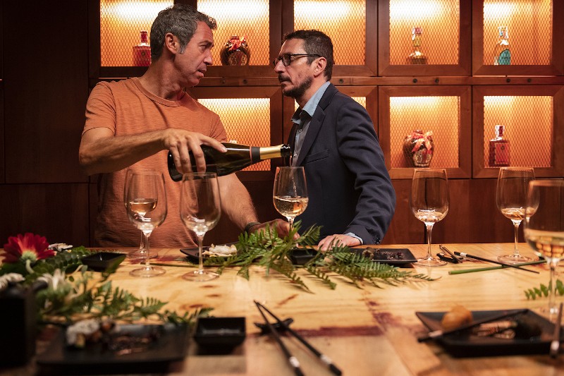 Dwóch mężczyzn stoi przy drewnianym stoliku barowym, na którym stoją puste kieliszki do wina, talerze do sushi i kwiaty. Jeden mężczyzna w koszulce nalewa wino, a drugi w garniturze przygląda się. Butelki są wystawione na półkach za nimi.