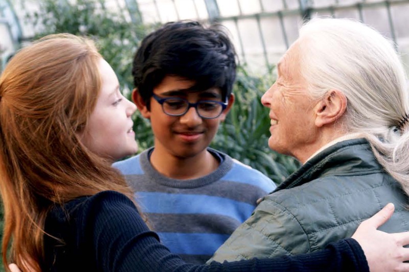 Starsza kobieta z białymi włosami przytula młodą dziewczynkę, podczas gdy chłopiec w okularach stoi obok i uśmiecha się. Są na zewnątrz, w tle jest zieleń, dzielą się ciepłym, radosnym momentem.