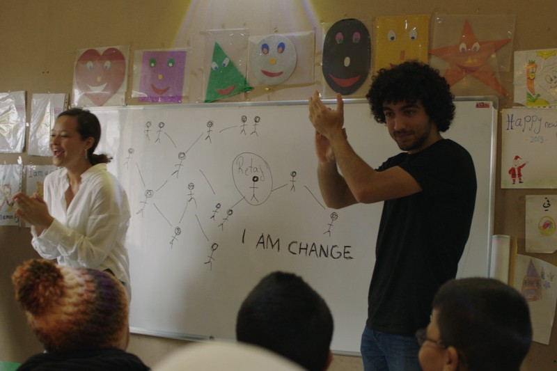 Dwie osoby stoją i klaszczą przed tablicą z rysunkami, słowem HOPE i frazą I AM CHANGE. Dzieci siedzą naprzeciwko nich. Na ścianie za tablicą wyświetlana jest kolorowa sztuka.