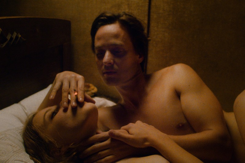 Mężczyzna i kobieta bez koszuli leżą razem w łóżku. Kobieta pali papierosa, podczas gdy mężczyzna delikatnie dotyka jej szyi i patrzy na nią. Oświetlenie jest ciepłe i intymne.