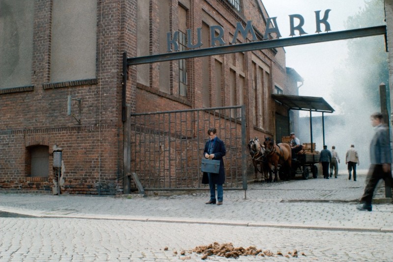 Mężczyzna w niebieskim mundurze stoi pod metalową bramą z napisem Kurmark na zewnątrz ceglanego budynku. W pobliżu stoi wóz konny i kilka osób; na brukowanej ulicy na pierwszym planie widać koński nawóz.
