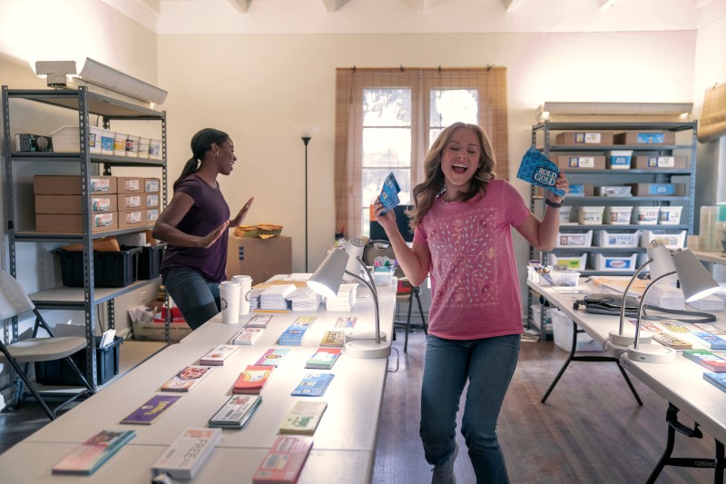 Dwie kobiety znajdują się w jasnym, uporządkowanym pomieszczeniu wypełnionym półkami do przechowywania i zapasami. Jedna kobieta tańczy radośnie, trzymając paczki, a druga układa przedmioty na stole pokrytym kolorowymi produktami.