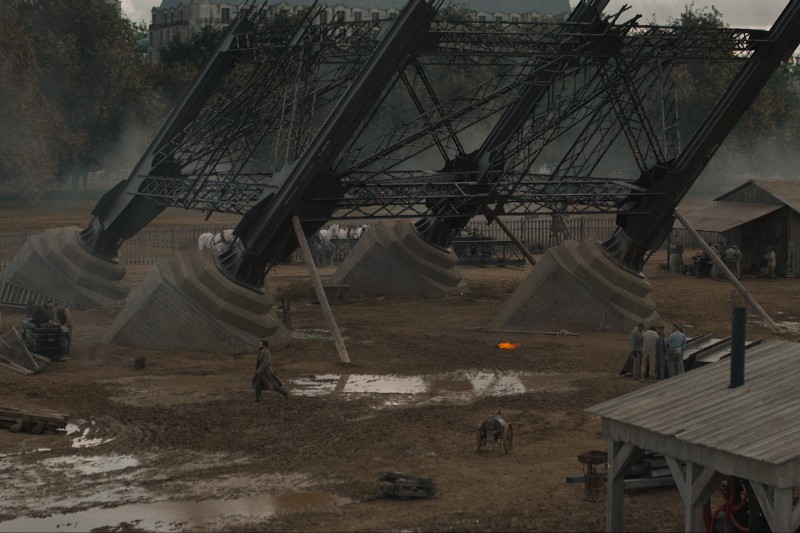 Częściowo zbudowane metalowe nogi Wieży Eiffla wyrastają z błotnistego gruntu, w otoczeniu robotników, drewnianych konstrukcji i materiałów budowlanych w pochmurny dzień.