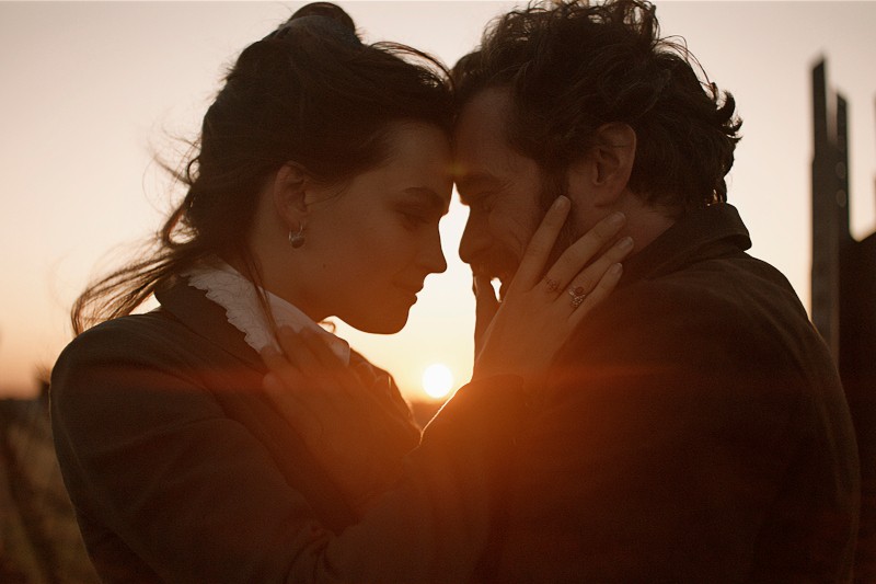 Para stoi blisko siebie o zachodzie słońca, delikatnie dotykając się czołami i trzymając się za twarze, na tle ciepłego światła słonecznego.