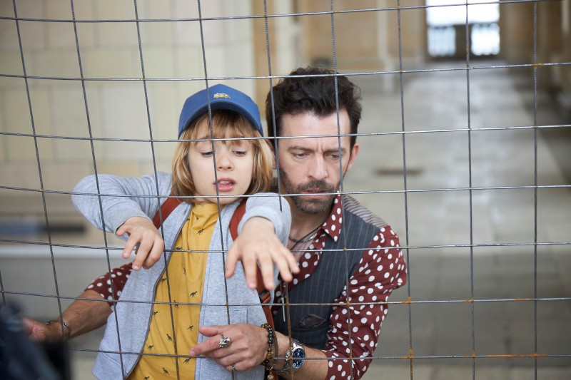 Mężczyzna i małe dziecko stoją za drucianym ogrodzeniem w pomieszczeniu. Dziecko, ubrane w niebieską czapkę i żółtą koszulkę, sięga przez ogrodzenie, podczas gdy mężczyzna, ubrany w koszulkę w kropki, podtrzymuje ich od tyłu.