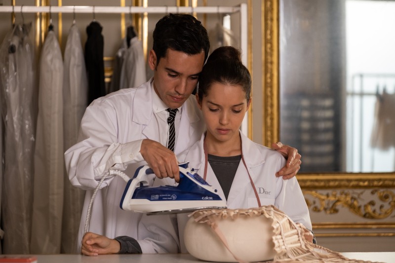 Mężczyzna i kobieta w białych fartuchach laboratoryjnych pracują razem, żelazkiem prasując ubranie w dobrze oświetlonym, eleganckim pomieszczeniu ze złotymi lustrami i ubraniami wiszącymi w tle.