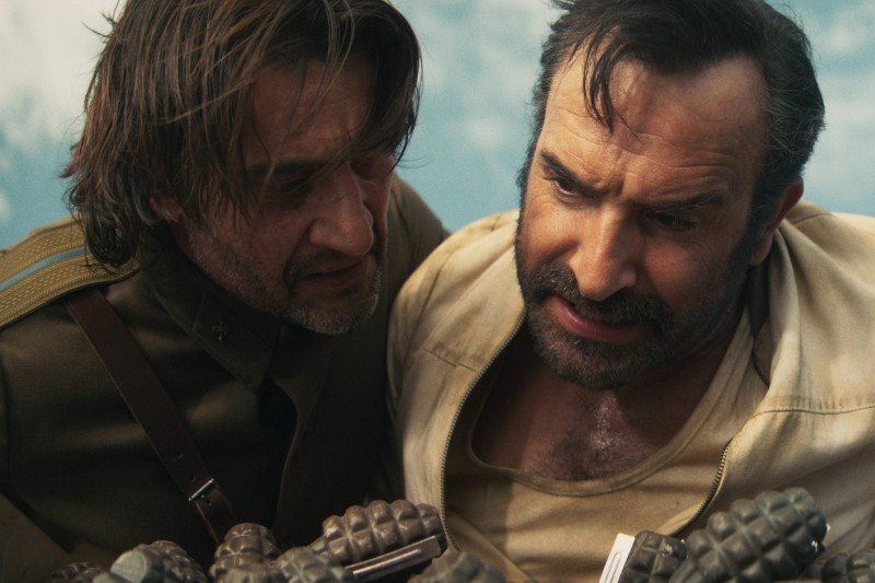 Dwóch mężczyzn, jeden w mundurze wojskowym, a drugi w jasnej koszuli, stoją blisko siebie i patrzą na kilka granatów ręcznych na pierwszym planie. Na ich twarzach maluje się zaniepokojenie.