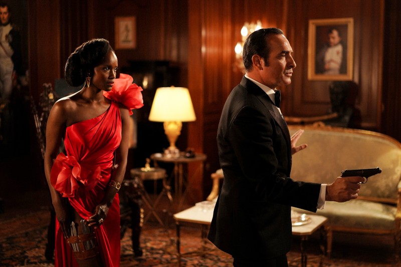 Kobieta w czerwonej sukience stoi czujnie z zabytkowym urządzeniem, podczas gdy mężczyzna w smokingu trzyma broń w bogato zdobionym, wyłożonym drewnem pokoju o klasycznym wystroju i z portretami na ścianach.