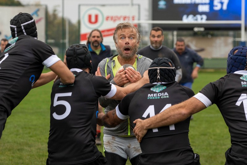 Zaskoczony gracz rugby trzyma piłkę mocno, gdy trzech członków drużyny przeciwnej w czarnych koszulkach rusza, aby go zaatakować podczas meczu na świeżym powietrzu. W tle widać widzów i tablicę wyników.