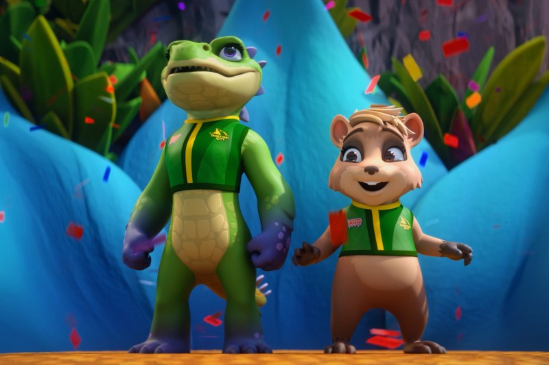 Dwie animowane postacie, zielona jaszczurka i brązowy wiewiór, ubrane w zielone kurtki, stoją dumnie na scenie, wokół której spadają niebieskie góry i kolorowe konfetti.