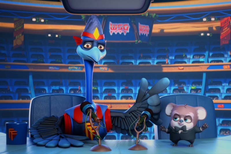 Niebieski struś w mundurze i mały koala w smokingu siedzą przy pulpicie komentatora z mikrofonami, na kolorowym stadionie z pustymi siedzeniami w tle.