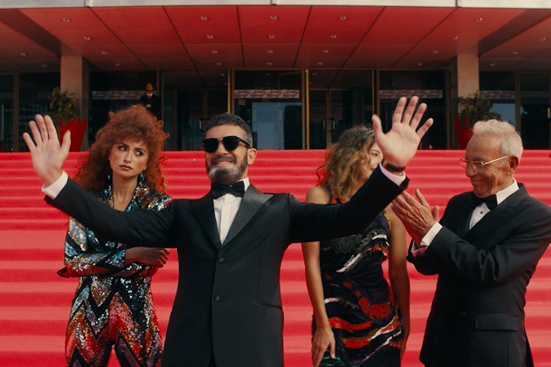 Mężczyzna w smokingu i okularach przeciwsłonecznych stoi na schodach wyłożonych czerwonym dywanem z uniesionymi ramionami, uśmiechając się pewnie. Trzy osoby za nim, formalnie ubrane, patrzą i klaszczą.