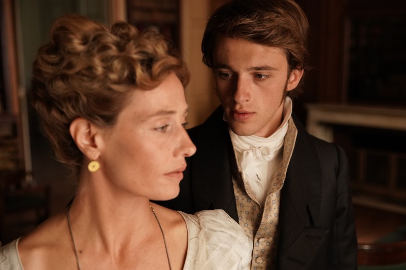 Kobieta w stroju z epoki odwraca wzrok, gdy młody mężczyzna w garniturze stoi za nią, patrząc na nią z poważnym wyrazem twarzy. Scenografia wydaje się historyczna, z ciepłym oświetleniem i zabytkowymi meblami.