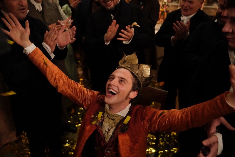 Młody mężczyzna w złotej koronie i pomarańczowej kurtce siedzi z uniesionymi ramionami, uśmiechając się radośnie, podczas gdy ludzie wokół niego klaszczą i świętują. Konfetti rozsypane jest na ziemi, tworząc świąteczną atmosferę.
