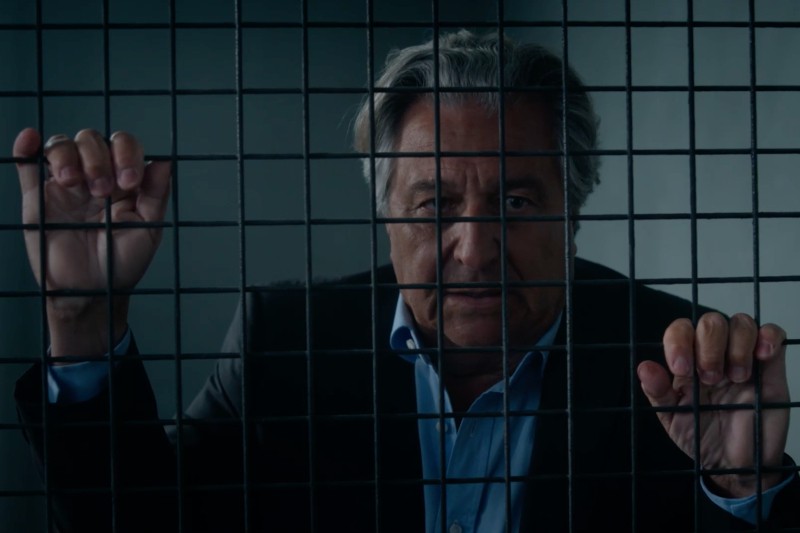 Starszy mężczyzna w garniturze ściska pręty metalowej kraty, uważnie przez nie patrząc. Oświetlenie jest słabe, a on wydaje się być w zamkniętej lub przypominającej więzienie przestrzeni.