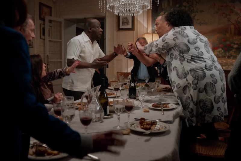 Przy stole wybucha ostra kłótnia, dwie osoby stoją i gestykulują ze złością, podczas gdy inni próbują interweniować. Na stole rozrzucone są talerze, szklanki i jedzenie.