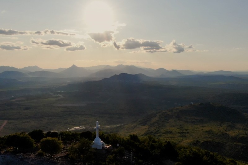 Jasne słońce świeci nad rozległym krajobrazem zielonych wzgórz i odległych gór. Na pierwszym planie widoczny jest biały pomnik, a na niebie rozproszone są chmury.