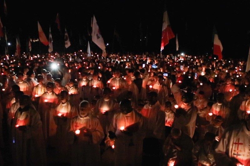 Duża grupa ludzi ubranych w białe szaty, wielu z czerwonymi krzyżami, trzyma zapalone świece podczas nocnego czuwania, otoczona flagami i oświetlona w ciemności blaskiem świec.