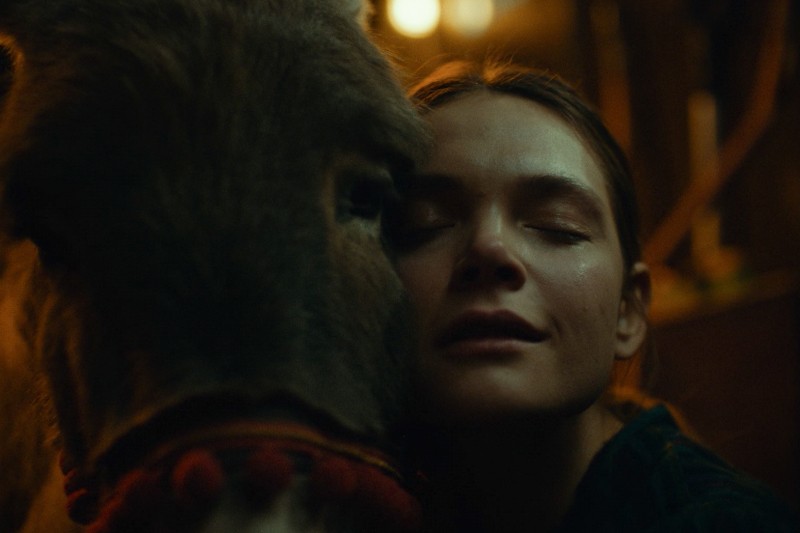 Kobieta z zamkniętymi oczami delikatnie opiera twarz o głowę osła w półmroku i ciepłym oświetleniu, co wyraża poczucie spokoju i więzi.