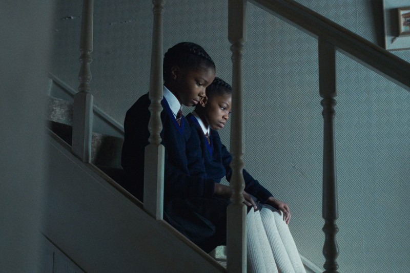 Dwoje dzieci w szkolnych mundurkach siedzi obok siebie na schodach, patrząc w dół z poważnymi minami. Oświetlenie jest słabe, co tworzy ponury i refleksyjny nastrój.
