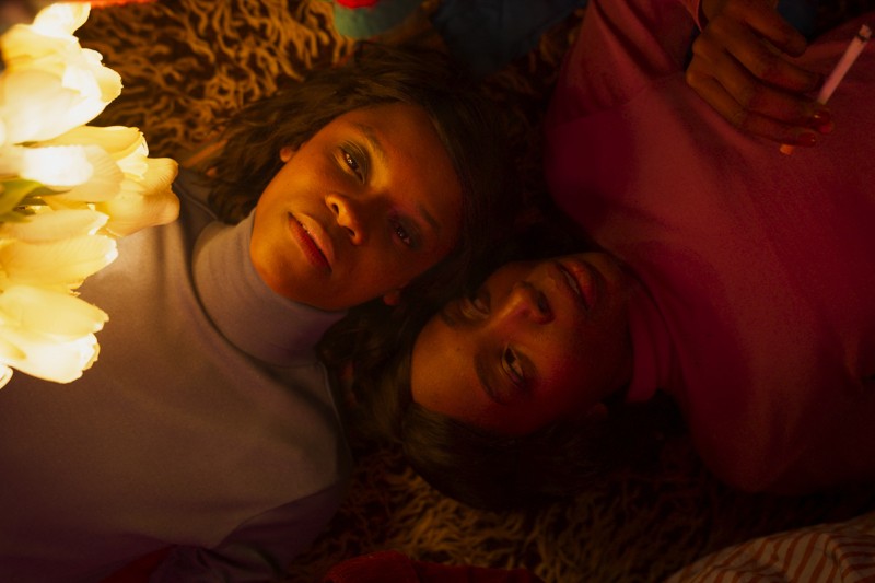 Dwie osoby leżą blisko siebie na teksturowanej powierzchni, oświetlone ciepłym światłem z pobliskich kwiatów. Jedna osoba ma na sobie lekki golf, druga różowy top i trzyma papierosa. Obie patrzą w górę ze spokojnym wyrazem twarzy.