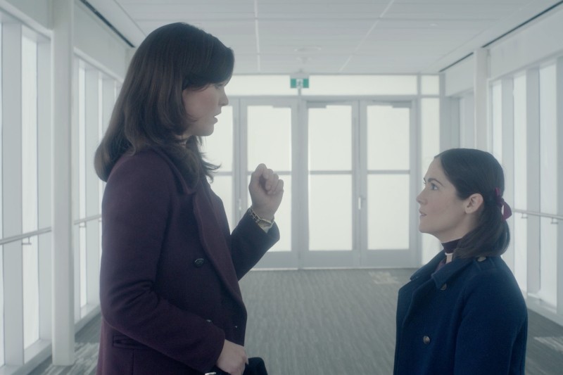 Dwie kobiety stoją naprzeciw siebie w jasnym korytarzu. Wyższa kobieta gestykuluje ręką, podczas gdy niższa kobieta patrzy na nią uważnie. Obie mają na sobie ciemne płaszcze.