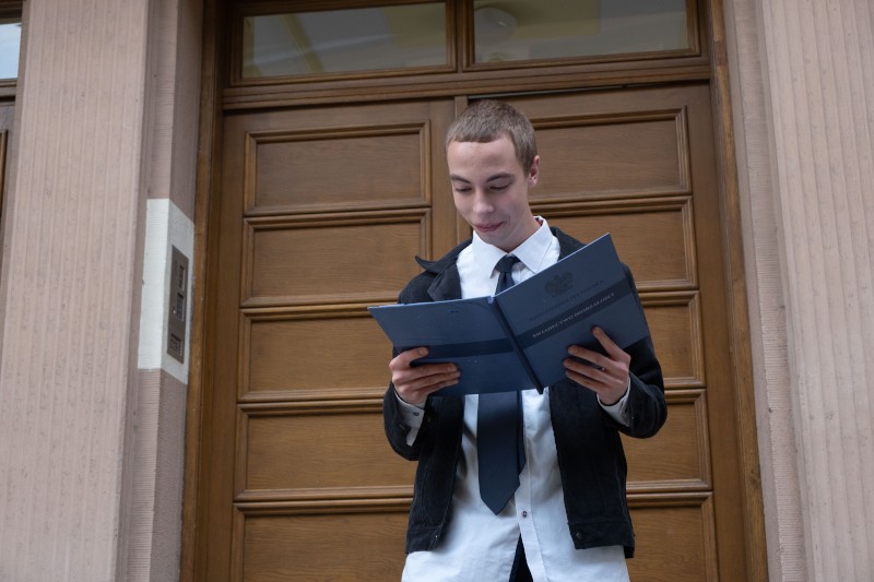 Młoda osoba ubrana w białą koszulę, czarny krawat i czarną marynarkę stoi przed drewnianymi drzwiami, patrzy i trzyma otwarty folder lub dokument.