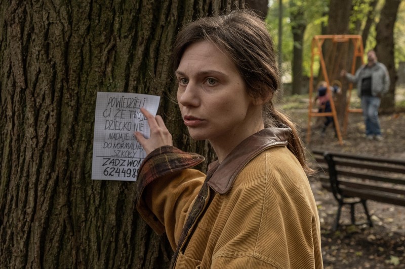 Kobieta w brązowej kurtce umieszcza ulotkę o zaginionej osobie na drzewie w parku, wyglądając na zaniepokojoną. W tle mężczyzna stoi przy pomarańczowej huśtawce i ławce wśród drzew.