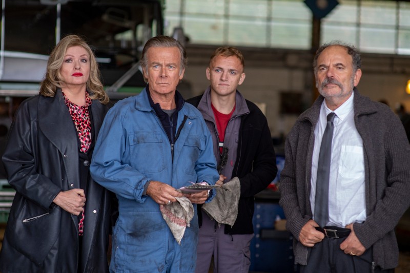 Czterech dorosłych stoi w garażu lub warsztacie. Jeden mężczyzna w niebieskim kombinezonie trzyma szmaty, obok niego kobieta w czarnym płaszczu i dwóch mężczyzn — jeden w swobodnym ubraniu, drugi w koszuli i krawacie. Wszyscy patrzą poważnie w kamerę.