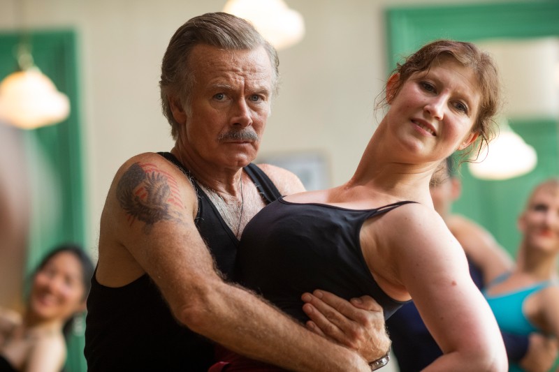 Starszy mężczyzna z wąsami i tatuażem trzyma uśmiechniętą kobietę w tanecznej pozie; oboje ubrani są w czarne stroje taneczne w jasno oświetlonym studiu z zielonymi ramami okien. Inni tancerze są rozmazani w tle.