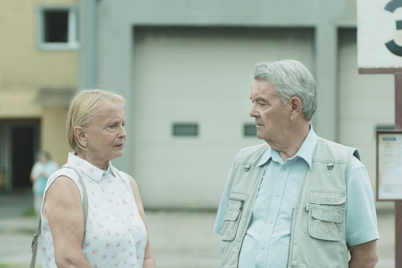 Starsza kobieta i mężczyzna stoją na zewnątrz, zwróceni do siebie i rozmawiający. W tle widać duży szary budynek i niewyraźną postać w oddali. Oboje wydają się spokojni i zaangażowani w rozmowę.