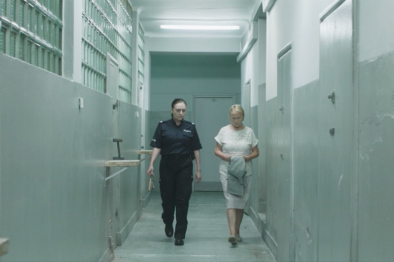 Strażnik więzienny eskortuje kobietę wąskim, szarym korytarzem więziennym z zakratowanymi oknami i zamkniętymi drzwiami celi. Kobieta patrzy w dół, trzymając złożoną odzież, podczas gdy strażnik idzie obok niej.