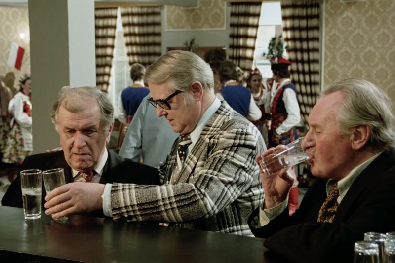 Trzech starszych mężczyzn w garniturach stoi przy barze, każdy trzymając szklankę przejrzystego płynu. Jeden mężczyzna w kraciastej marynarce sprawdza swój drink, podczas gdy pozostali dwaj piją. Ludzie w świątecznych, tradycyjnych strojach są w tle.