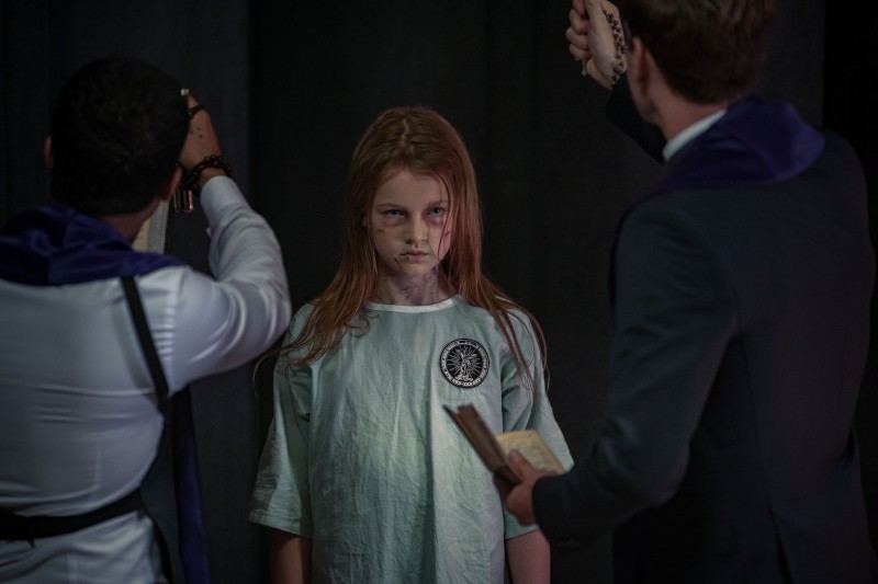 Młoda dziewczyna z potarganymi włosami i szpitalnym fartuchem stoi między dwoma mężczyznami trzymającymi przedmioty religijne, co sugeruje scenę egzorcyzmów. Dziewczyna patrzy do przodu z poważnym, intensywnym wyrazem twarzy.