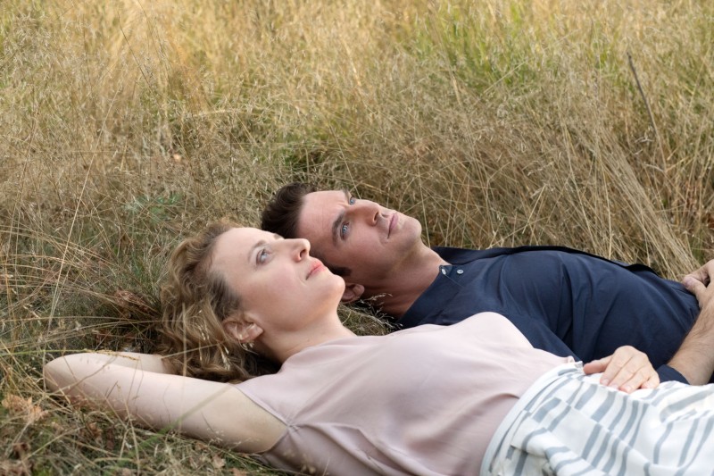 Kobieta i mężczyzna leżą obok siebie w wysokiej, suchej trawie, patrząc zamyśleni w górę. Kobieta ma kręcone blond włosy i nosi jasną bluzkę, podczas gdy mężczyzna ma ciemne włosy i nosi granatową koszulę.