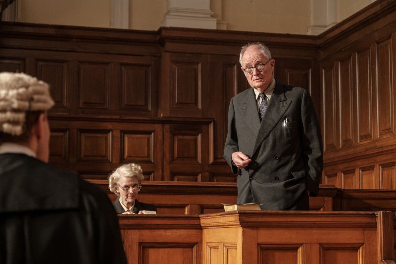 Starszy mężczyzna w garniturze stoi i przemawia w wyłożonej drewnem sali rozpraw, podczas gdy starsza kobieta siedzi w pobliżu i robi notatki, a na pierwszym planie widoczna jest osoba w białej peruce.