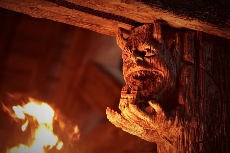 Zbliżenie drewnianej belki wyrzeźbionej z twarzą i ręką dzikiego, rogatego stworzenia, oświetlonej ciepłym blaskiem pobliskiego ognia. Tekstura drewna i płomienie tworzą dramatyczną, rustykalną atmosferę.