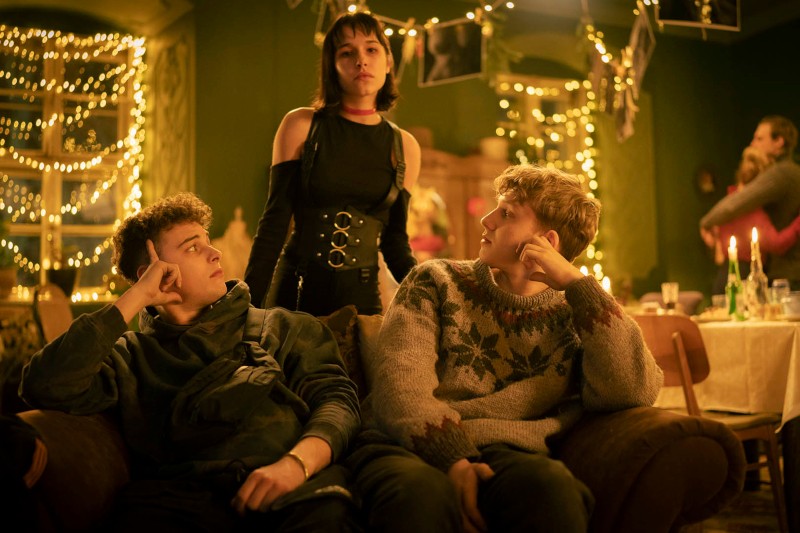 Trzech młodych dorosłych znajduje się w ciepło oświetlonym pokoju, udekorowanym lampkami i zdjęciami. Dwóch siedzi na kanapie, patrząc w górę na trzecią osobę, która stoi i patrzy na nich z góry. Inni mieszają się w tle.