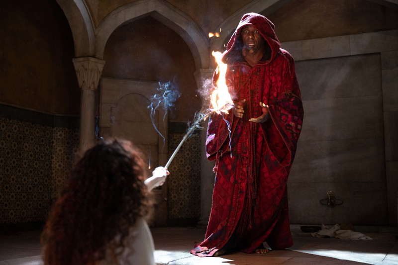 Osoba w czerwonej szacie z kapturem stoi boso w pomieszczeniu, trzymając ręce w górze, gdy ogień rozbłyska z laski skierowanej w jej stronę przez inną osobę klęczącą na pierwszym planie. W pomieszczeniu znajdują się łukowate drzwi i wzorzyste kafelki.