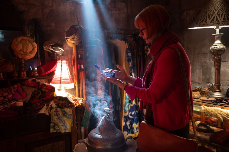 Kobieta w słabo oświetlonym, eklektycznym pokoju bada niebieską szklaną butelkę, podczas gdy dym unosi się z pobliskiego naczynia. Światło słoneczne wpada przez okno, oświetlając ją i kolorowe tkaniny wokół niej.