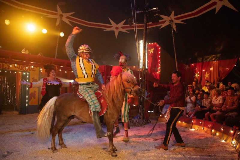 Scena cyrkowa przedstawia klauna jadącego na kucyku, podnoszącego jedną rękę w powietrze. Inni wykonawcy i klauni stoją w pobliżu, a publiczność siedzi w tle pod jasnymi światłami i świątecznymi dekoracjami.