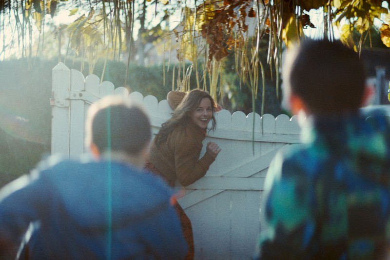 Kobieta w kostiumie uśmiecha się, bawiąc się przy białym płocie, spoglądając na dwójkę dzieci w kolorowych kurtkach biegnących w jej stronę na zewnątrz w słoneczny dzień.