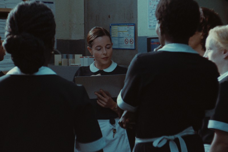 Kobieta w stroju pokojówki trzyma podkładkę i przemawia do grupy pokojówek, które stoją wokół niej w słabo oświetlonym pokoju i uważnie słuchają.