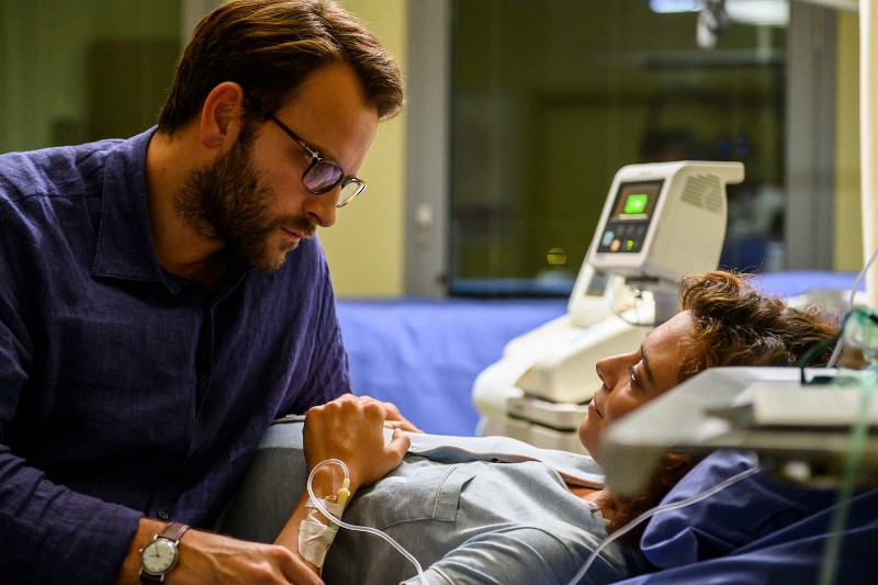 Mężczyzna w okularach delikatnie trzyma rękę kobiety leżącej na szpitalnym łóżku z kroplówką, podczas gdy patrzą na siebie z troską. Obok łóżka widać sprzęt medyczny i monitory.