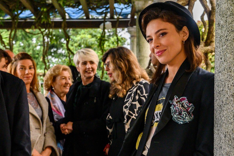 Kobieta w ciemnej marynarce i kapeluszu uśmiecha się do kamery na pierwszym planie, podczas gdy grupa kobiet stoi razem i rozmawia w tle pod zewnętrznym zadaszeniem otoczonym zielenią.