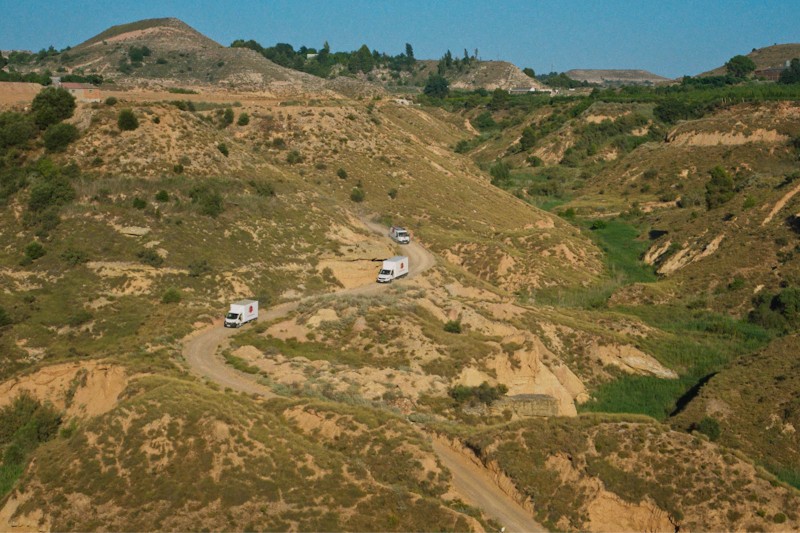 Trzy białe ciężarówki jadą krętą, gruntową drogą przez surowy, pagórkowaty krajobraz z suchą trawą i kępami zielonej roślinności pod czystym, błękitnym niebem.