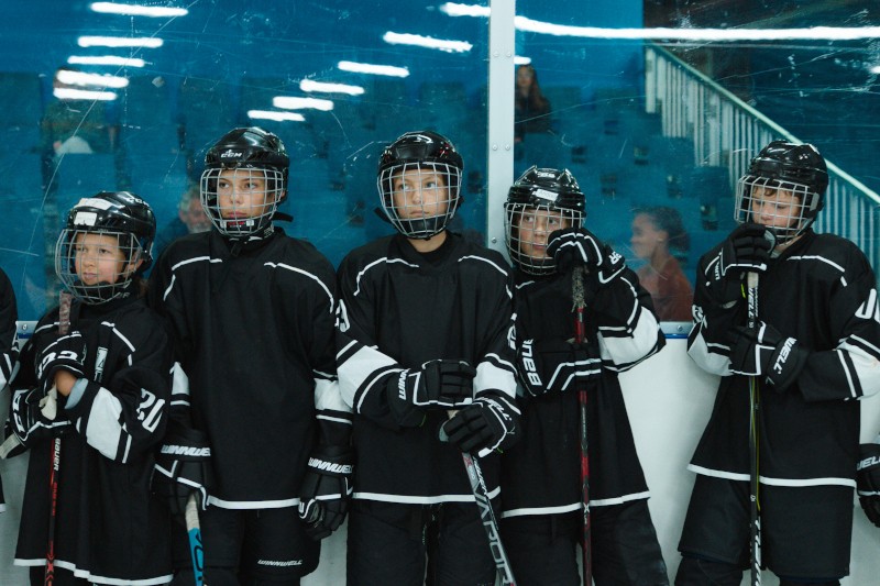 Pięciu młodych hokeistów w czarnych strojach i kaskach stoi obok siebie na lodowisku, trzymając kije hokejowe. W tle widać niebieską ścianę i siedzących widzów.