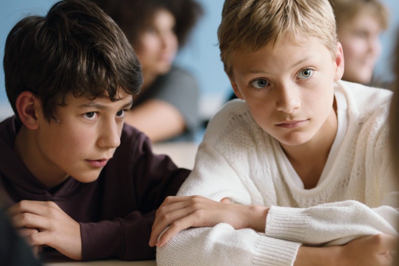 Dwóch chłopców siedzi blisko siebie przy biurku w klasie, uważnie słuchając. Jeden z nich ma na sobie ciemną bluzę z kapturem, drugi biały sweter. Obaj wydają się skupieni, z niewyraźnymi kolegami z klasy w tle.