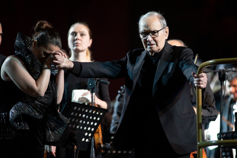 Starszy mężczyzna w czerni podnosi rękę, trzymając dłoń kobiety, która wydaje się być wzruszona. Muzycy z instrumentami stoją za nimi na scenie, jedna kobieta patrzy przed siebie z powagą.