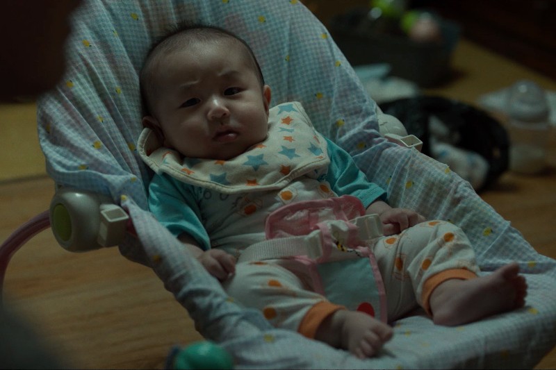 Dziecko ubrane w wzorzystą piżamę i śliniaczek siedzi w wyściełanym foteliku dziecięcym, wyglądając na lekko zdezorientowane lub zaniepokojone. Sceneria wydaje się być w pomieszczeniu z delikatnym oświetleniem.